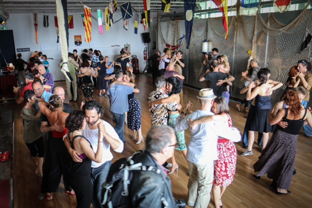 Color photo of an indoor social dance venue. It captures approximately thirty couples, dressed informally, in various tango gestures and embraces. Above the wooden dance floor hang numerous international flags and banners