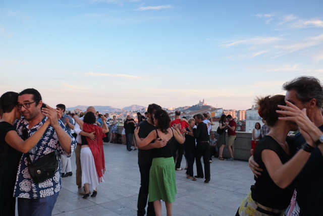 Color photo of an outdoor social dance venue. It captures approximately ten couples, dressed informally, dancing on a pavilion in various tango gestures and embraces in the foreground. The evening sky illuminates the background of a natural hilly landscape, with an old church or fortress on one hill, and more modern apartment buildings in the middle ground.  