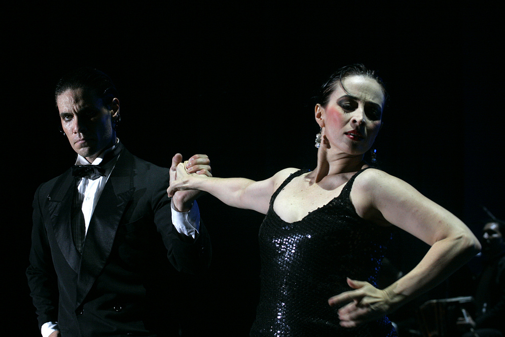 Color photo of a tango show-dance couple, dressed in formal black evening attire. As they clasp hands, the woman looks away from her outstretched arm, while the man gazes intently at her. 