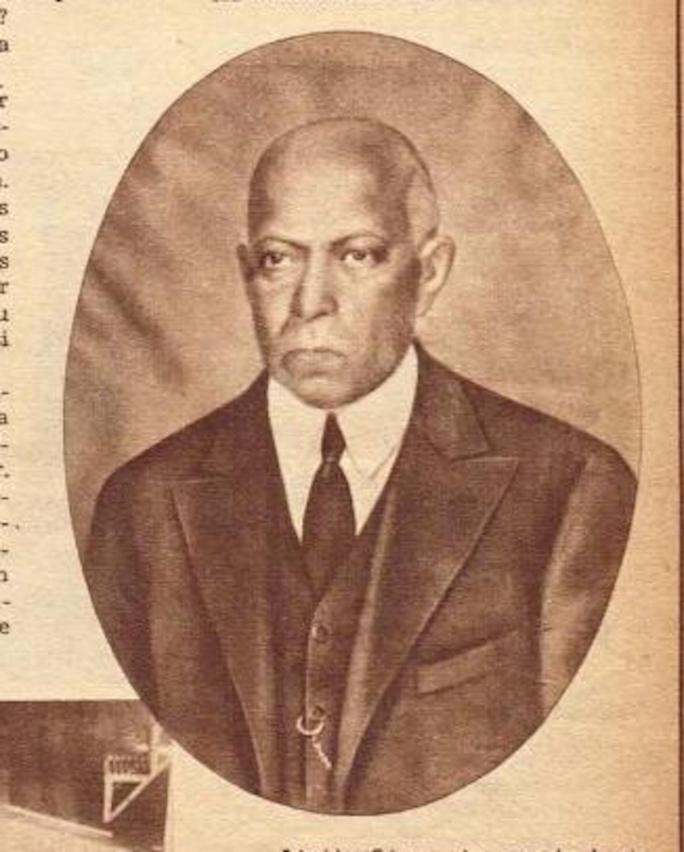 Archival sepia photo of a distinguished elder Black man from torso to head. He is finely dressed in a suit, vest, and tie, and his serious facial expression is somewhat severe.