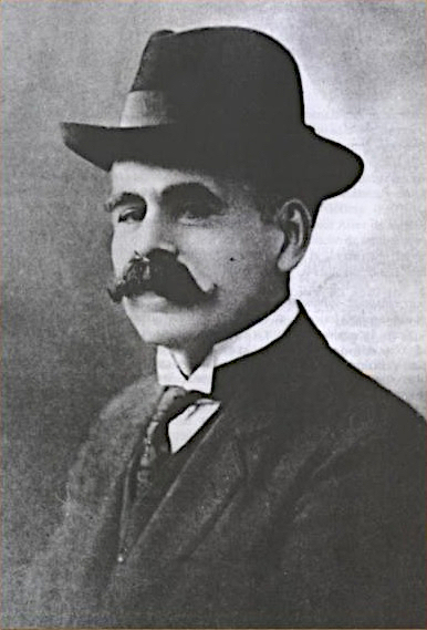 Black and white photo of a man in a dark suit and hat. He appears to be in his twenties or thirties and has a curved mustache.
