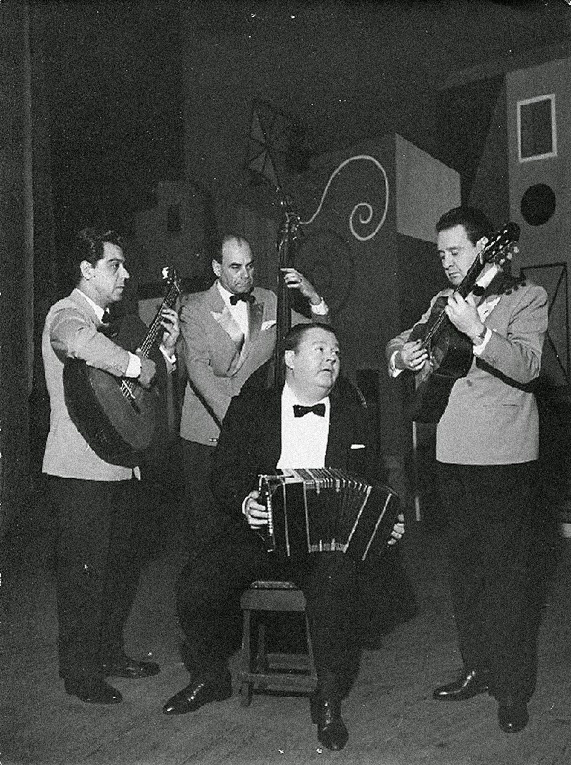 Black and white photo of a quartet. Aníbal Troilo is in a tuxedo, sitting center, looking right, and playing the bandoneón. Roberto Grela is in a tuxedo with a light jacket, standing on the right, and playing the guitar. Ernesto Báez is in a tuxedo with a light jacket, standing on the left, looking at Grela, and playing the guitarrón. Eugenio Pro is in a tuxedo with a light jacket, standing behind Troilo and playing the bass.