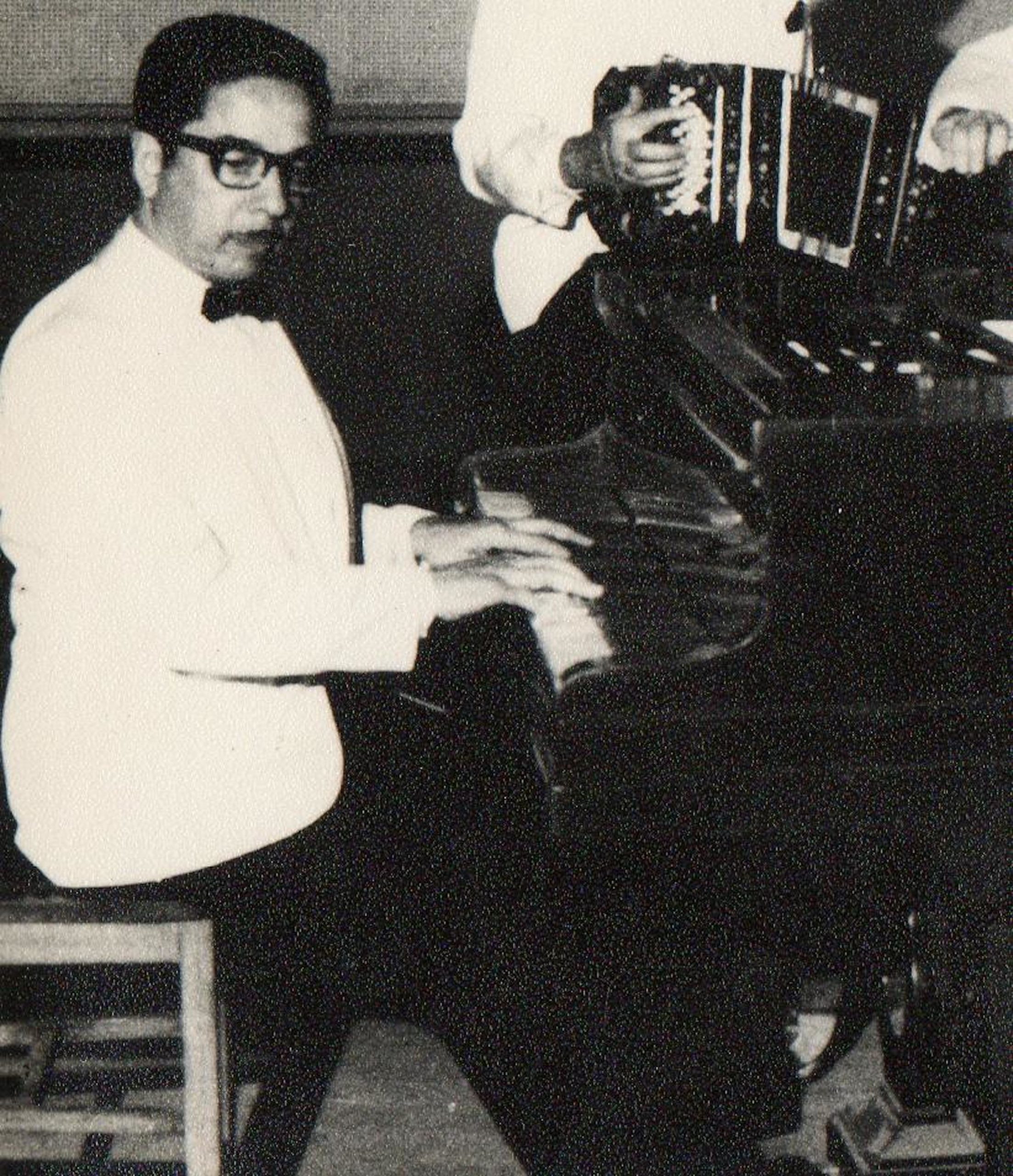 A black and white photo of Horacio Salgán sitting at the piano. He is wearing a tuxedo with a white jacket. A person in a white jacket playing the bandoneón is in the background behind the piano.