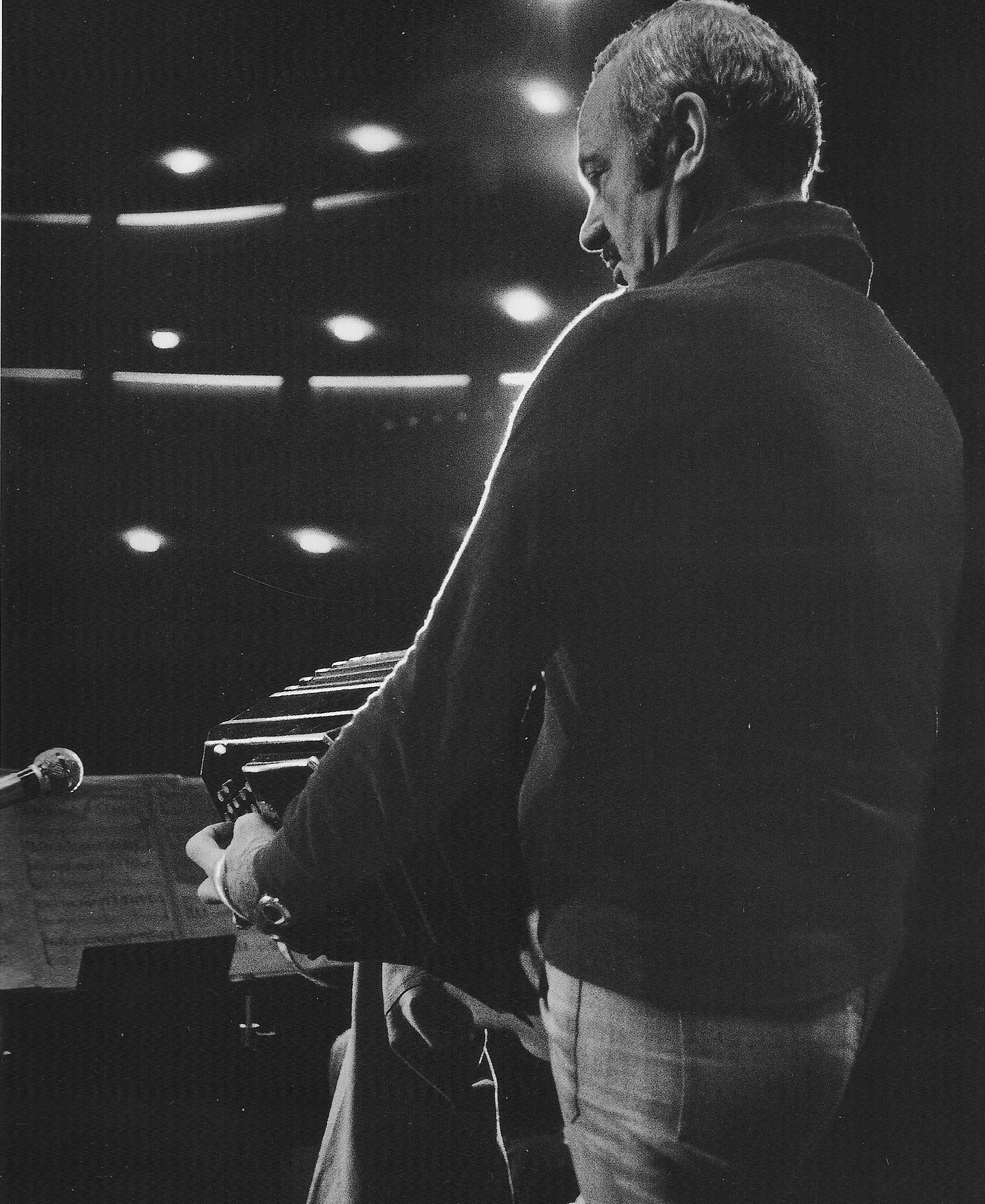 A black and white photo of Astor Piazzolla standing on a stage playing the bandoneón. The photo is taken from behind Piazzolla and there is a view of his music stand and the stage lights. He is wearing jeans and a sweater.